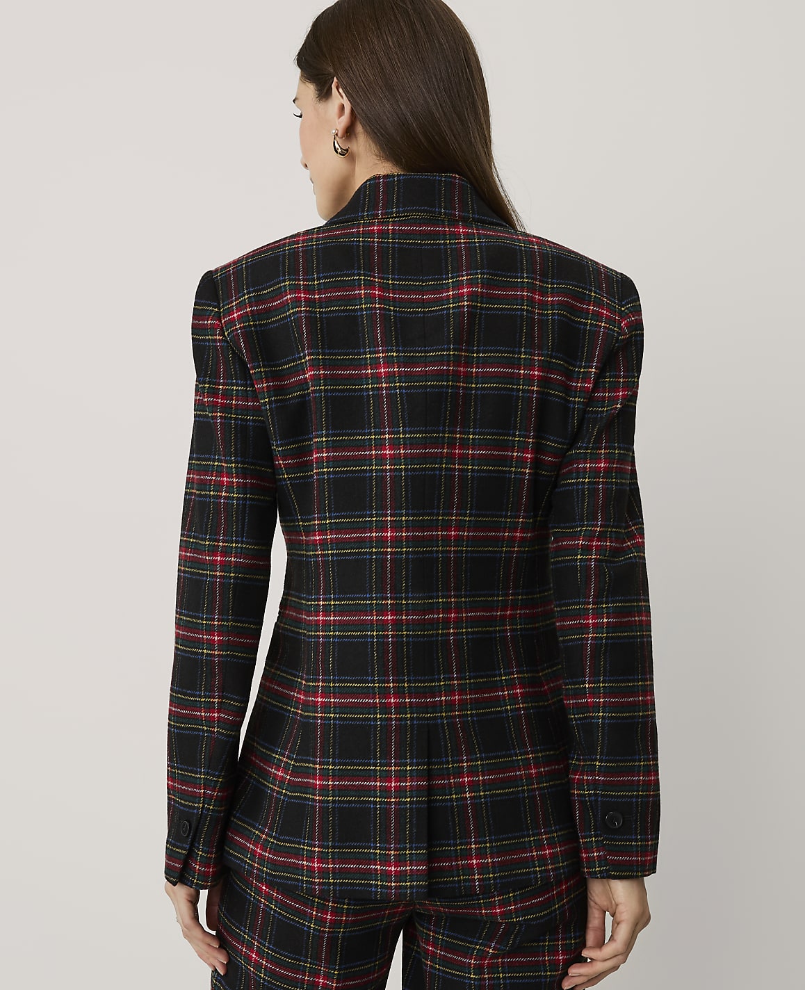 A woman stands with her back to the camera wearing a red, yellow, and blue plaid blazer and matching trousers against a plain light grey background.