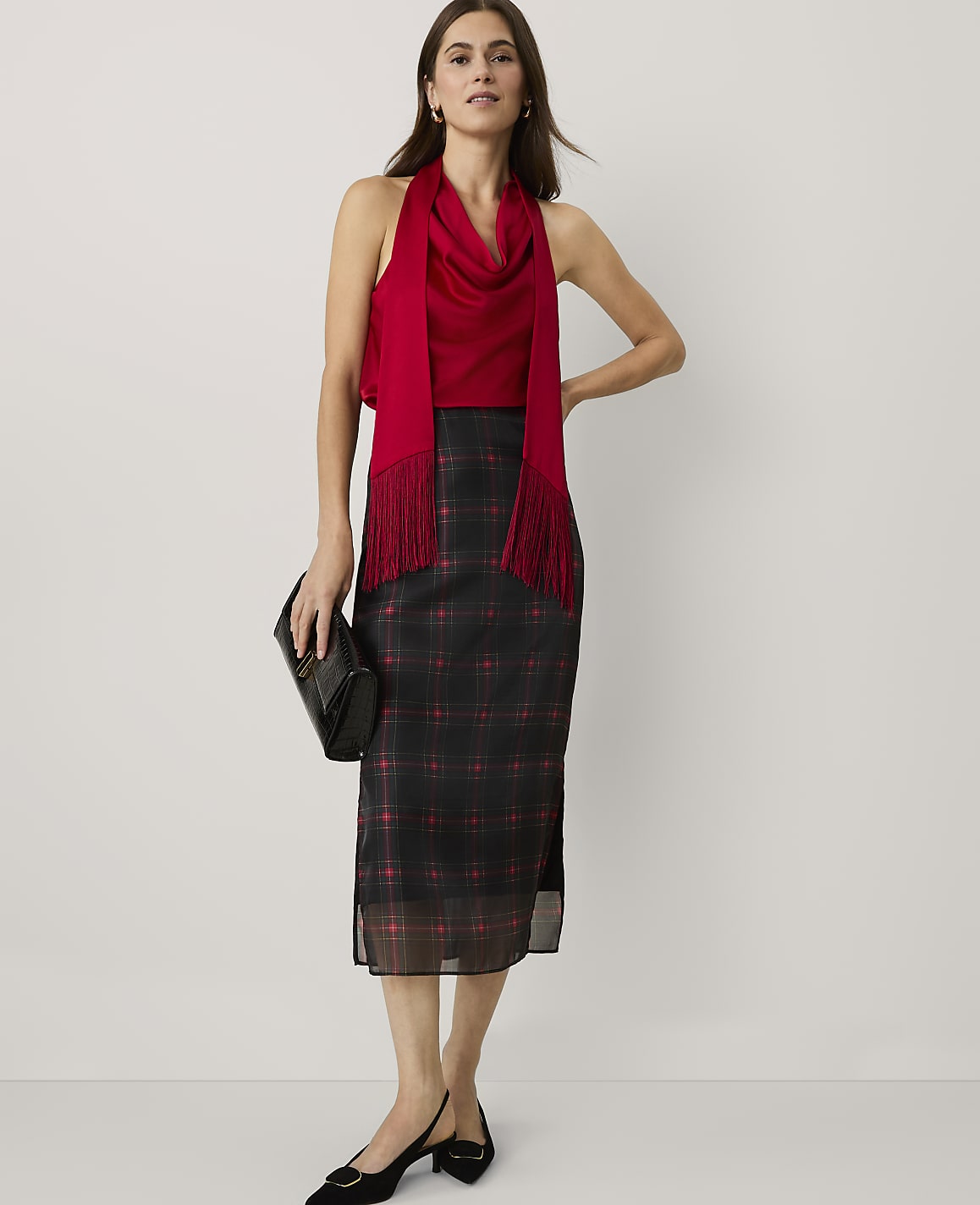 A woman wearing a red top and a black plaid skirt stands against a plain white background.