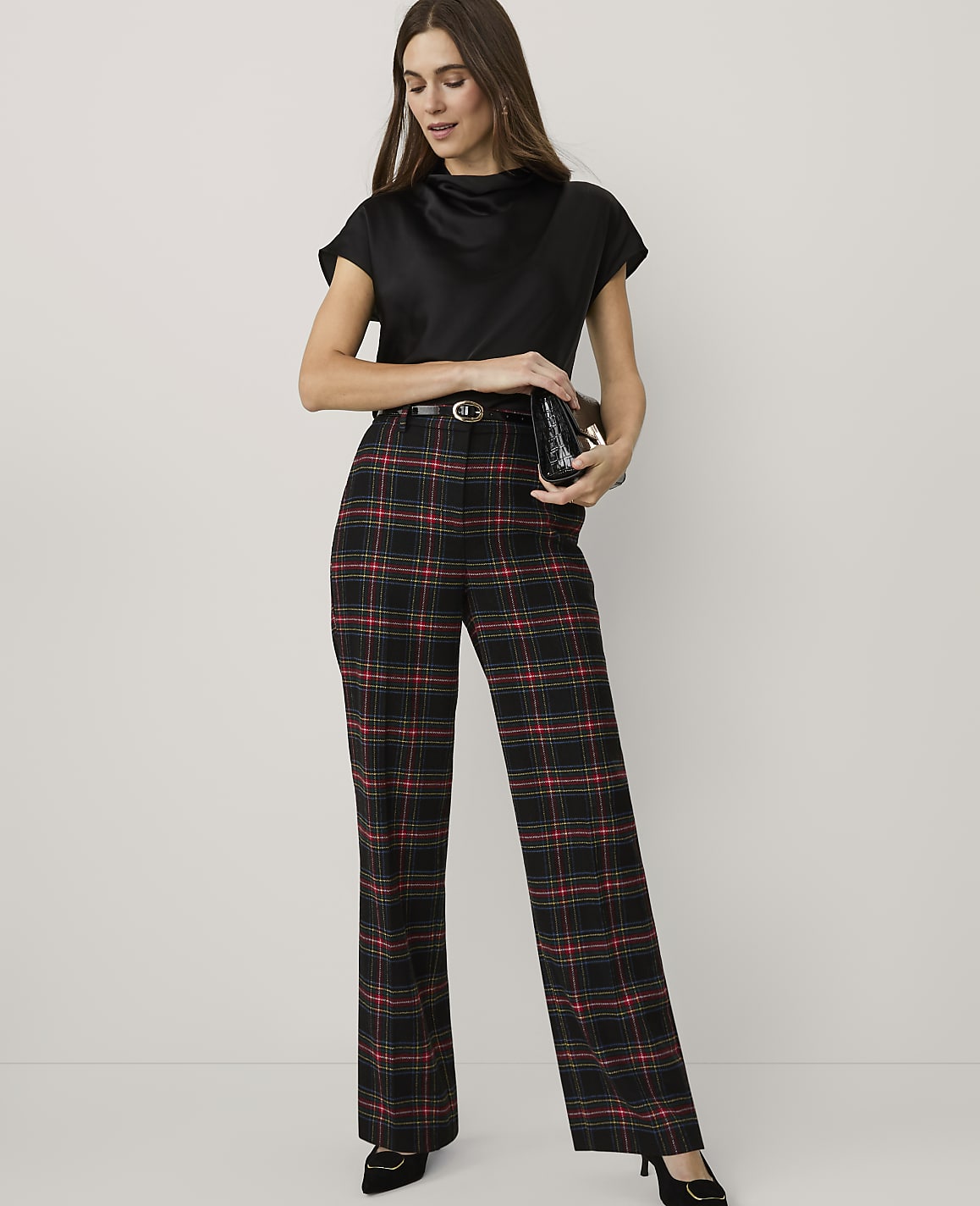 A woman wearing a black top and red and black plaid trousers stands against a plain white background.