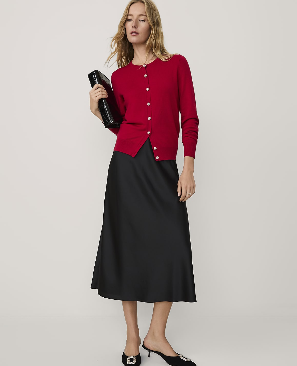 A woman wearing a red cardigan and a black midi skirt stands against a plain white background.