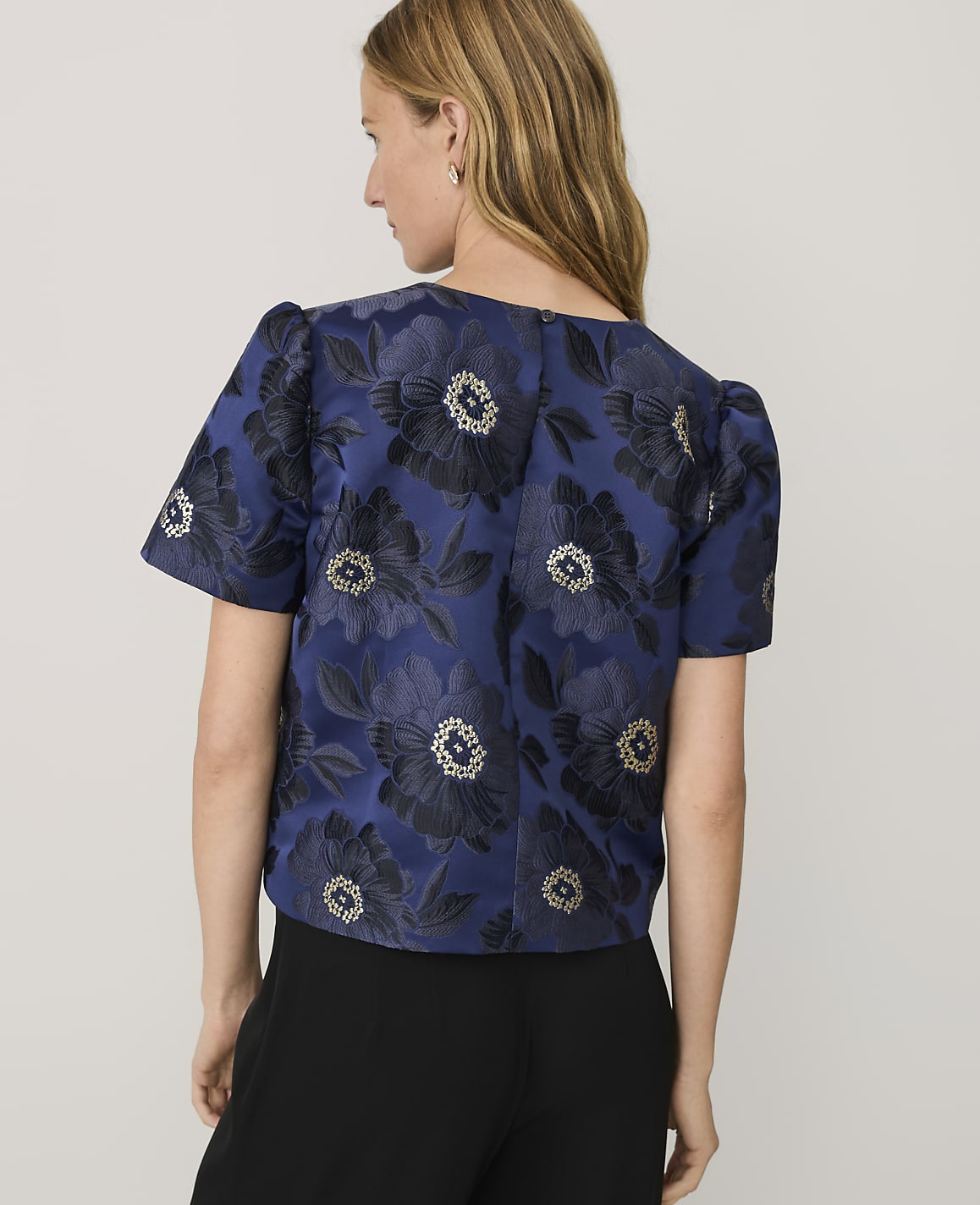 A woman wearing a blue floral jacquard top and black trousers stands against a plain light gray background.