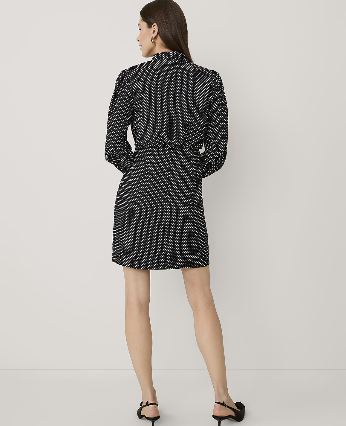 A woman stands with her back to the camera wearing a black and white polka dot dress and black heels against a plain white background.