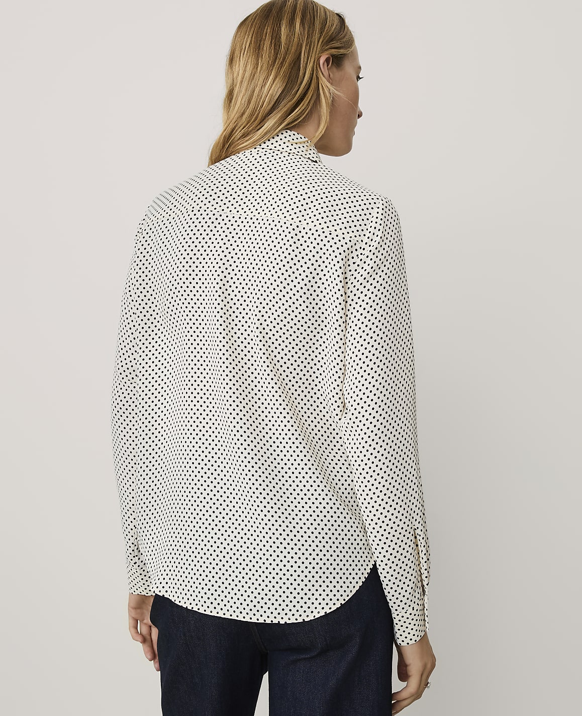 A woman wearing a white polka-dot shirt and dark blue pants stands against a plain light gray background.