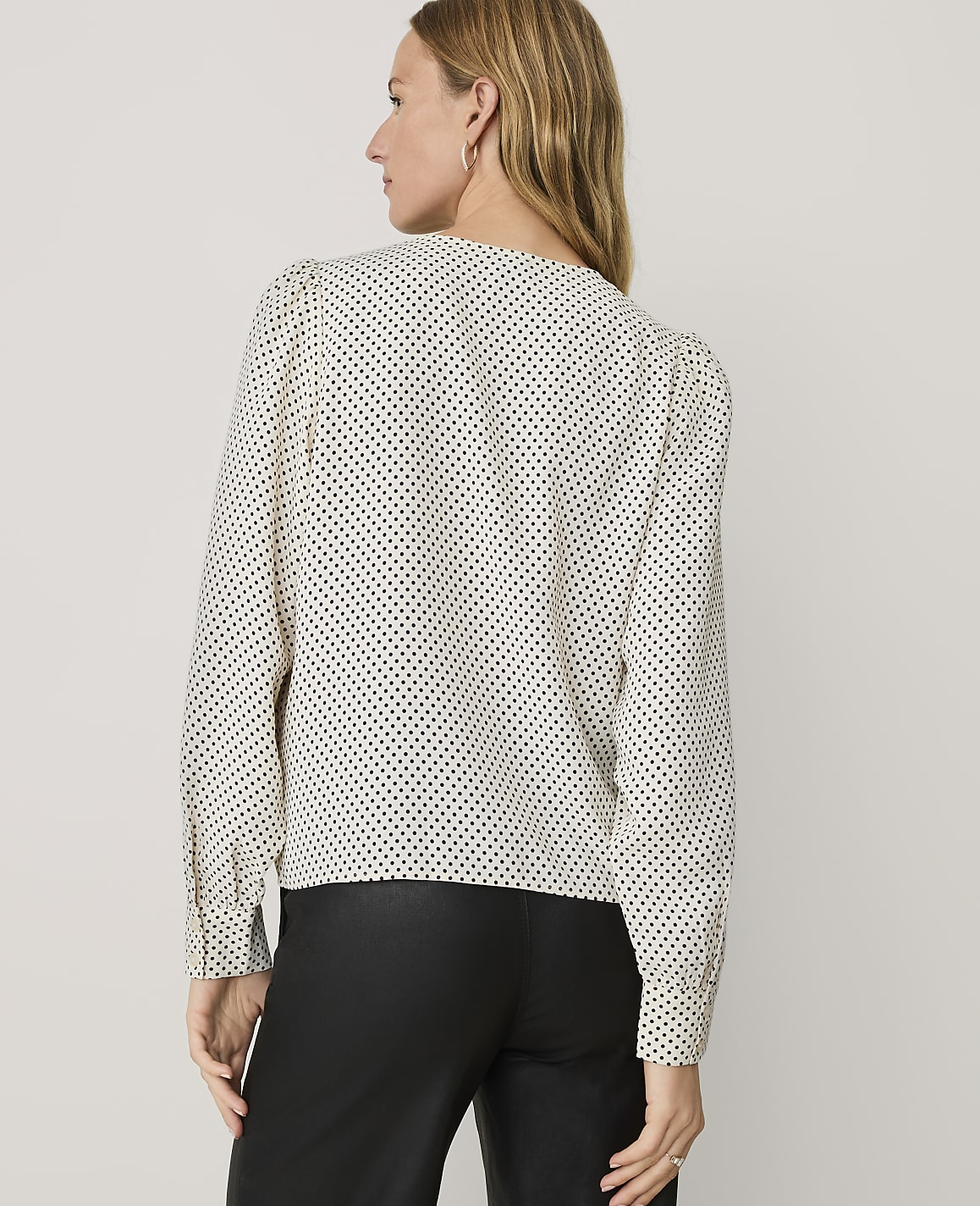 A woman wearing a white polka-dot blouse and black trousers stands with her back to the camera against a plain light gray background.
