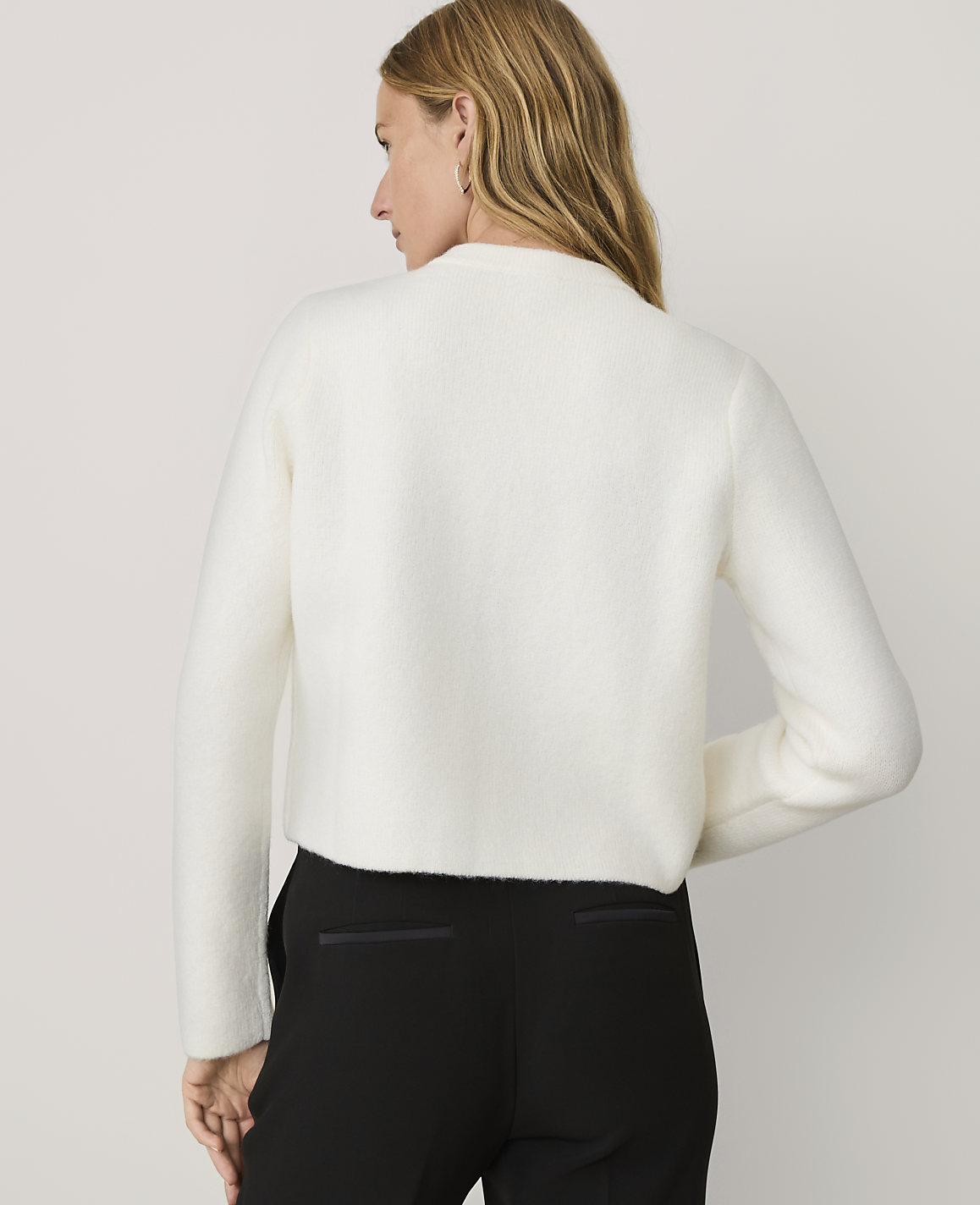A woman wearing a white fuzzy sweater and black trousers stands against a plain light gray background.