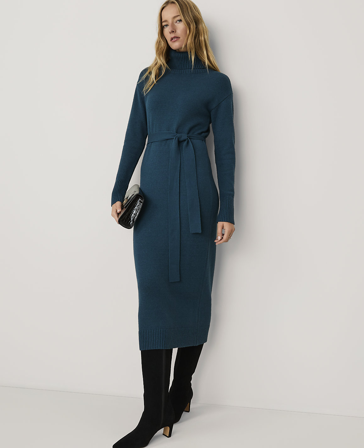 A woman wearing a long teal knit dress and black boots stands against a plain white background.