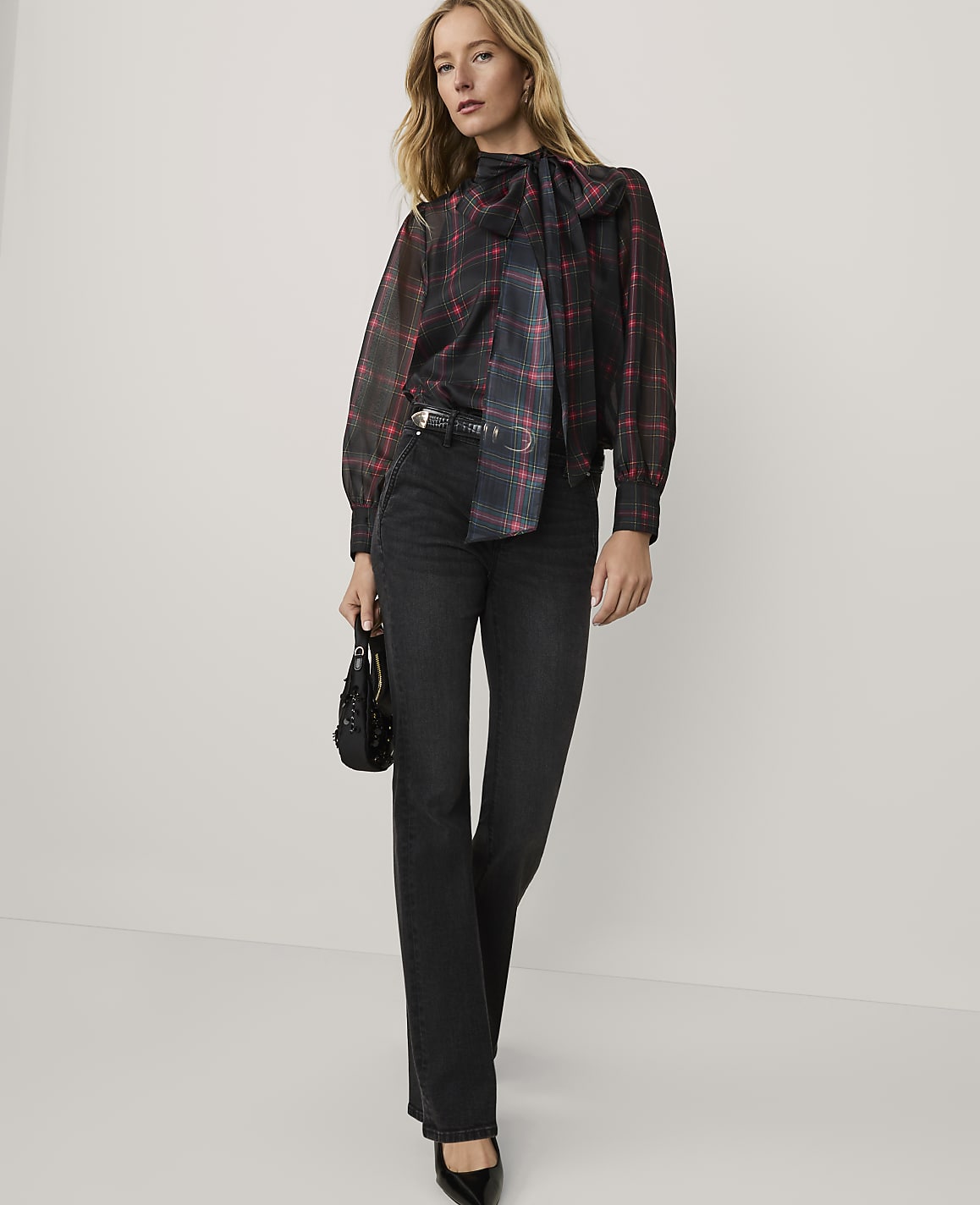 A woman wearing a red and black plaid bow-neck blouse and black jeans stands against a plain white background.