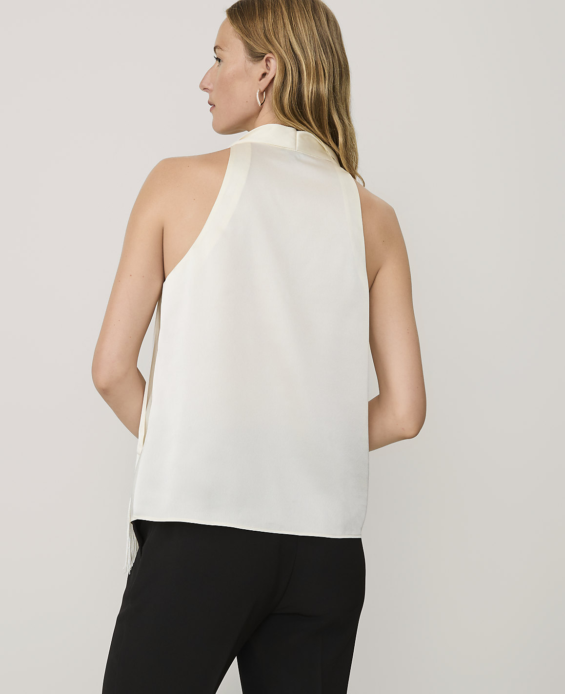 A woman wearing a white sleeveless top and black trousers stands against a plain light gray background.