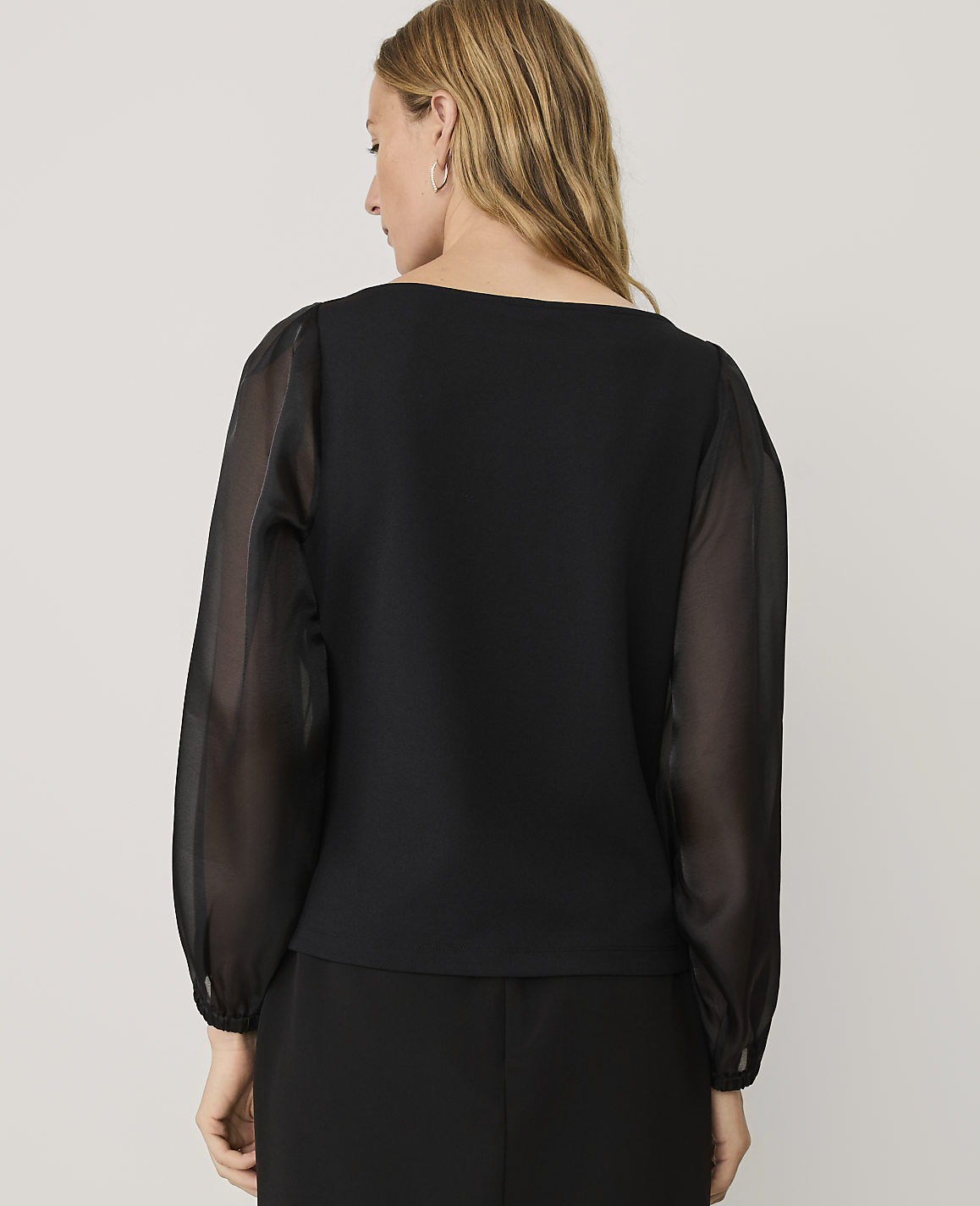 A woman wearing a black top with sheer sleeves stands with her back to the camera against a plain light gray background.