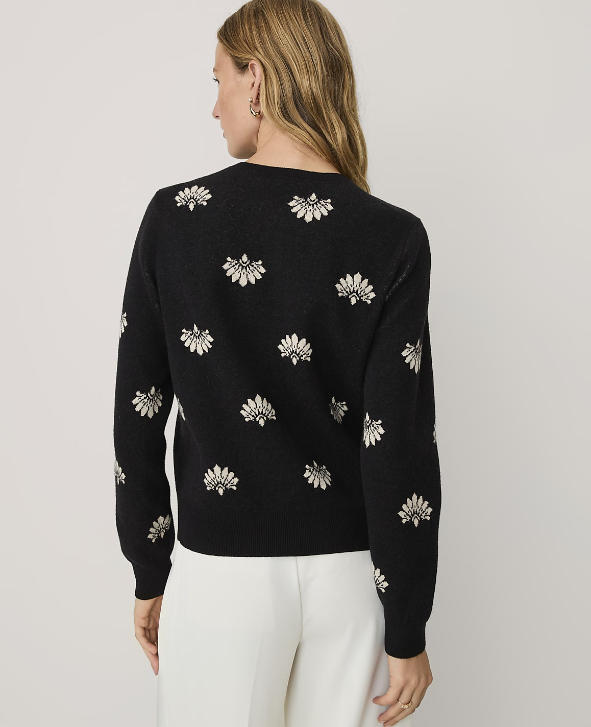 A woman wearing a black floral-patterned sweater and white trousers stands against a plain light gray background.