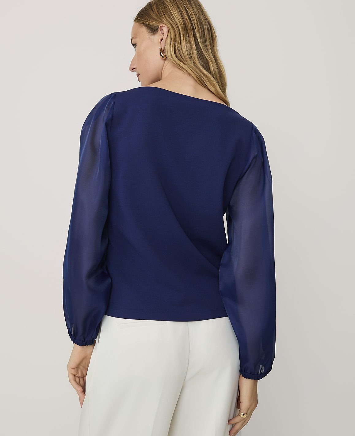 A woman wearing a navy blue blouse and white trousers stands against a plain light gray background.