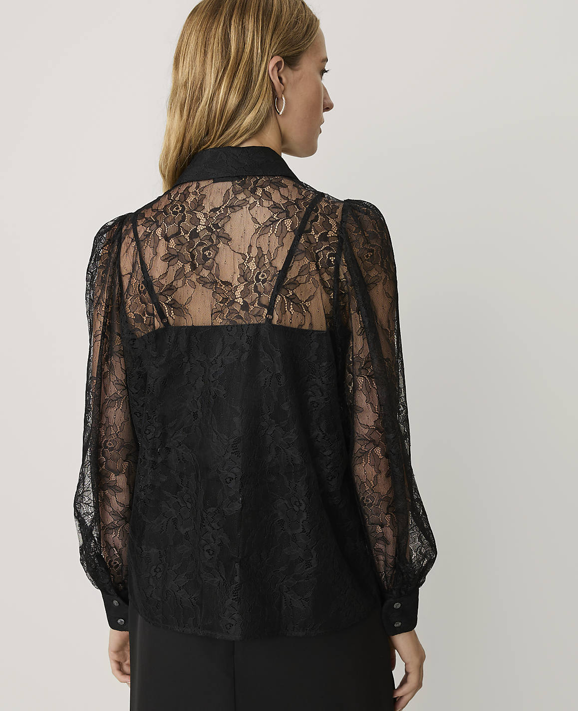 A woman wearing a black lace blouse stands with her back to the camera against a plain white background.