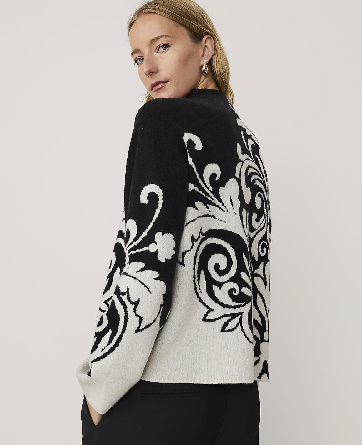 A woman wearing a black and white patterned sweater and black pants stands against a plain, light-colored background.