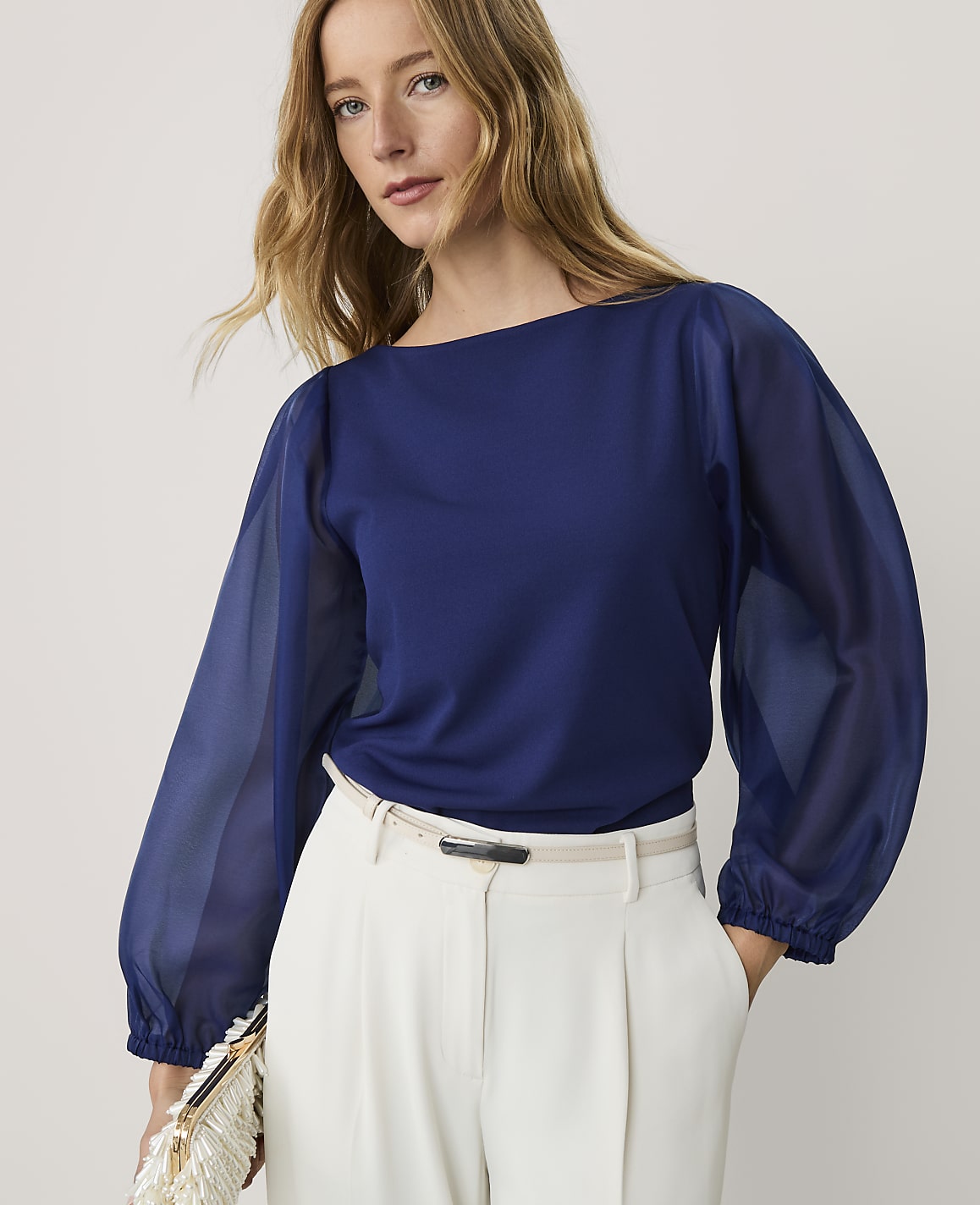 A woman wearing a navy blue blouse and white trousers stands against a plain, light-colored background.