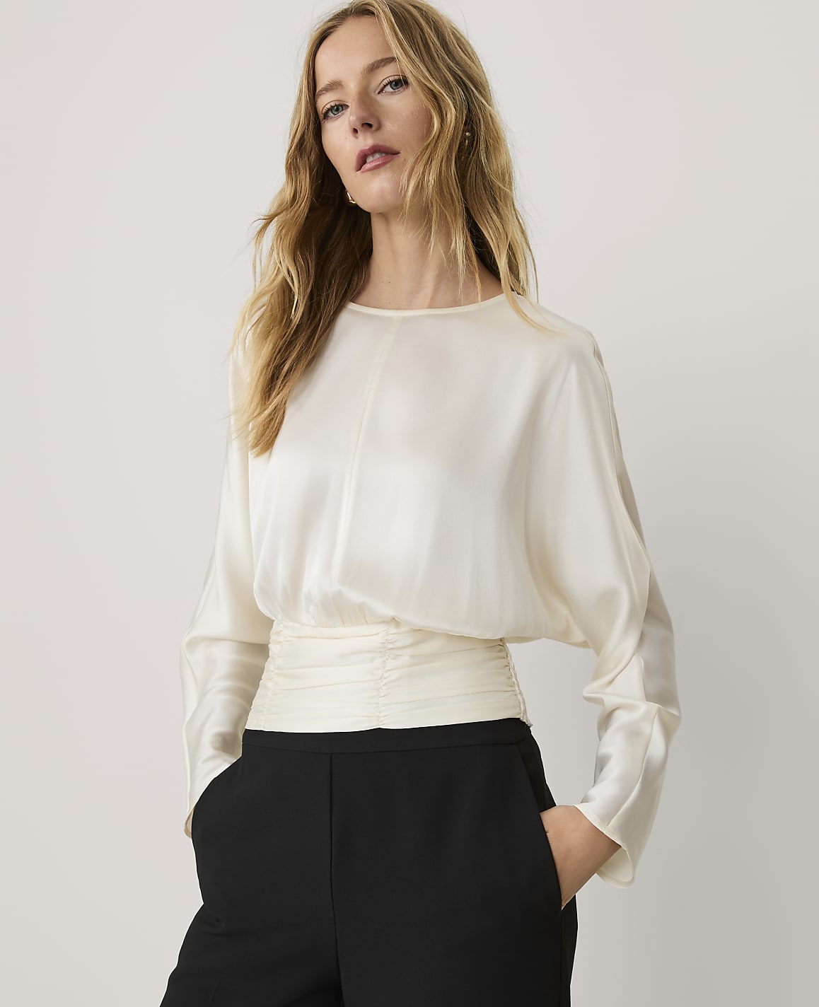 A woman wearing a cream-colored satin blouse and black trousers stands against a plain, light-colored background.