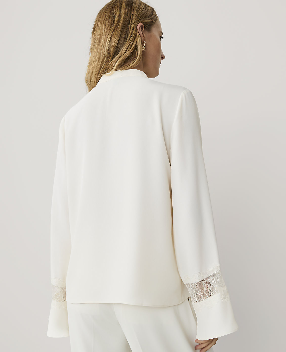 A woman with blonde hair is seen from behind wearing a white satin jacket with lace cuffs against a plain light grey background.