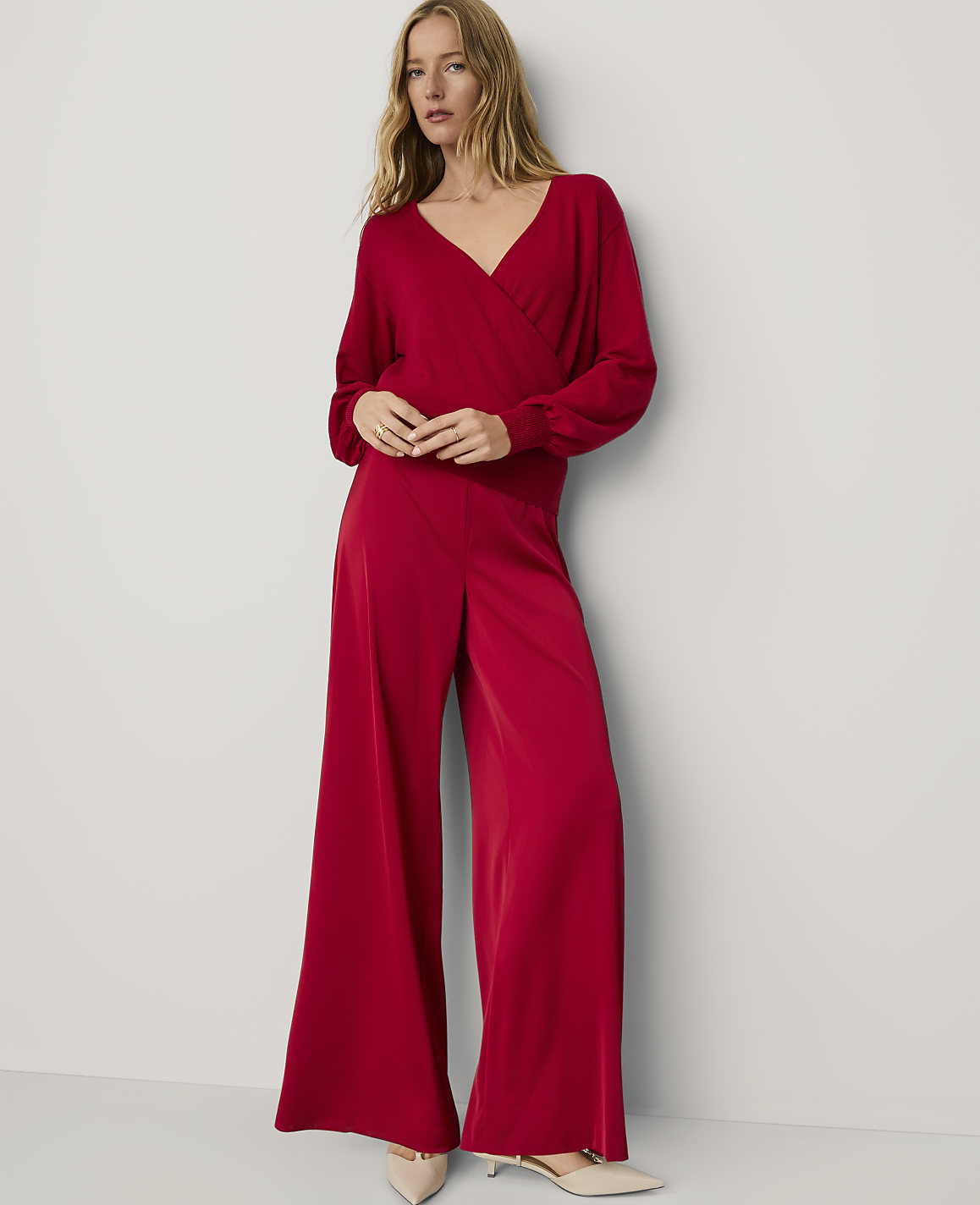 A woman wearing a red wrap-style jumpsuit and beige pointed-toe heels stands against a plain white wall.