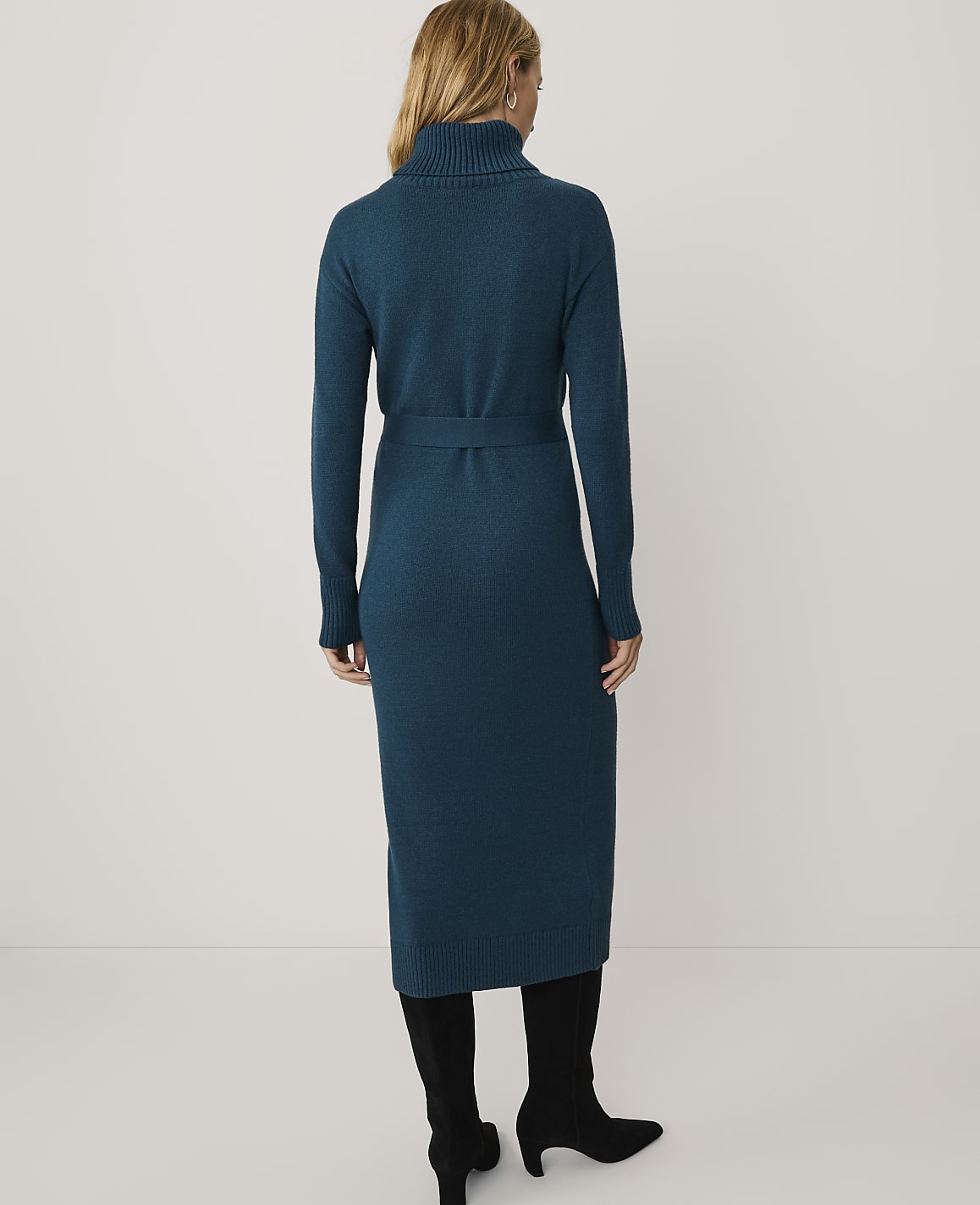 A woman stands with her back to the camera wearing a long-sleeved teal turtleneck knit dress and black boots against a plain white background.
