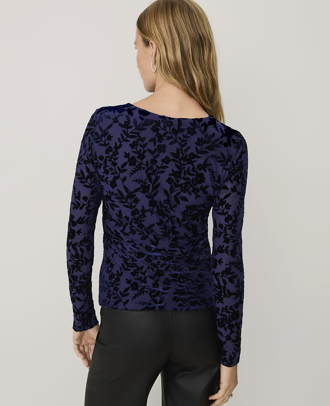 A woman wearing a navy blue floral velvet top and black leather pants stands against a plain light gray background.