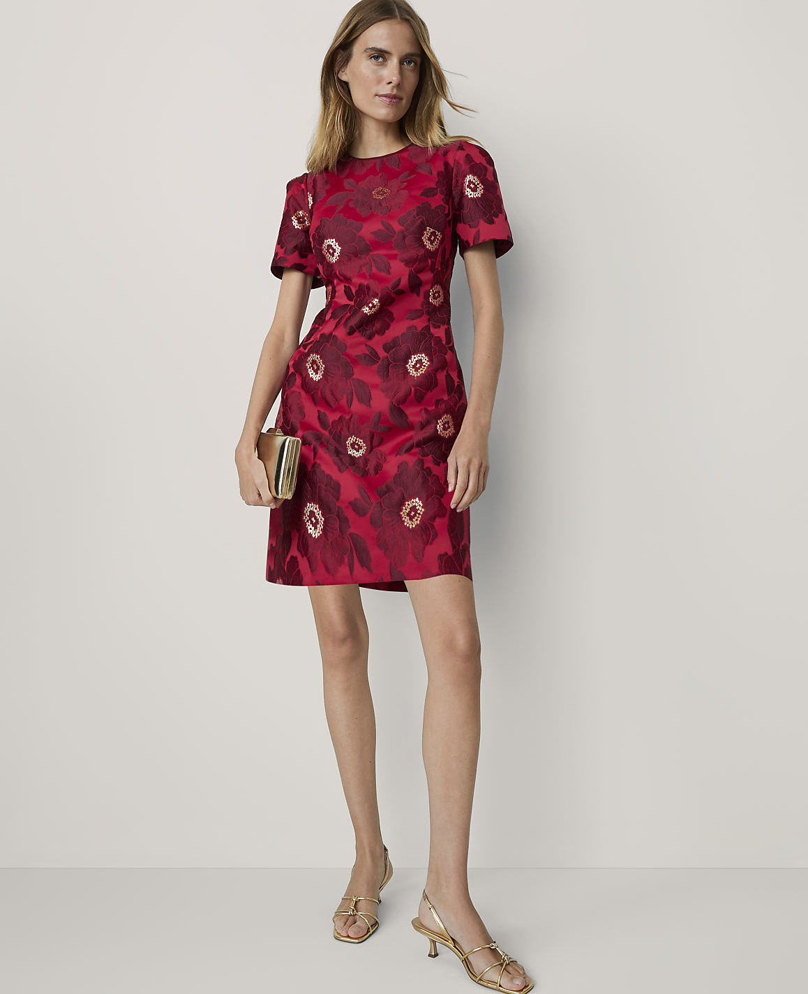 A woman in a red floral dress and gold heels stands against a plain white background, holding a gold clutch.