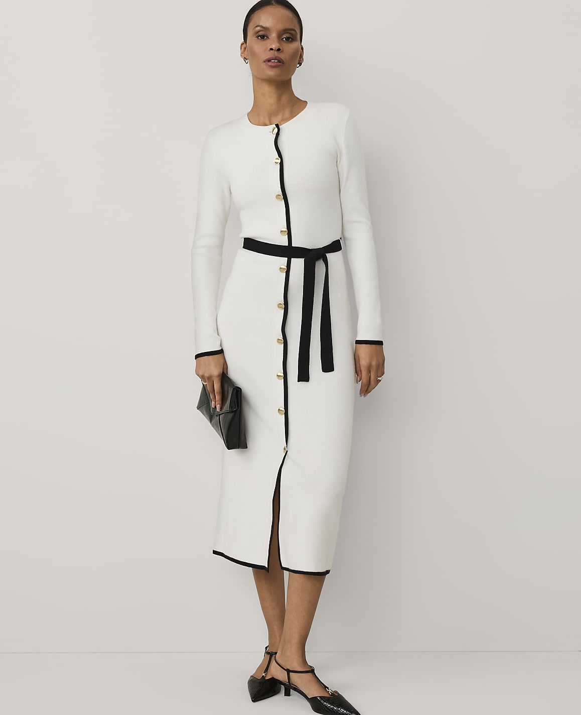 A woman wearing a white long-sleeved dress with black trim and a black belt stands against a plain white background.