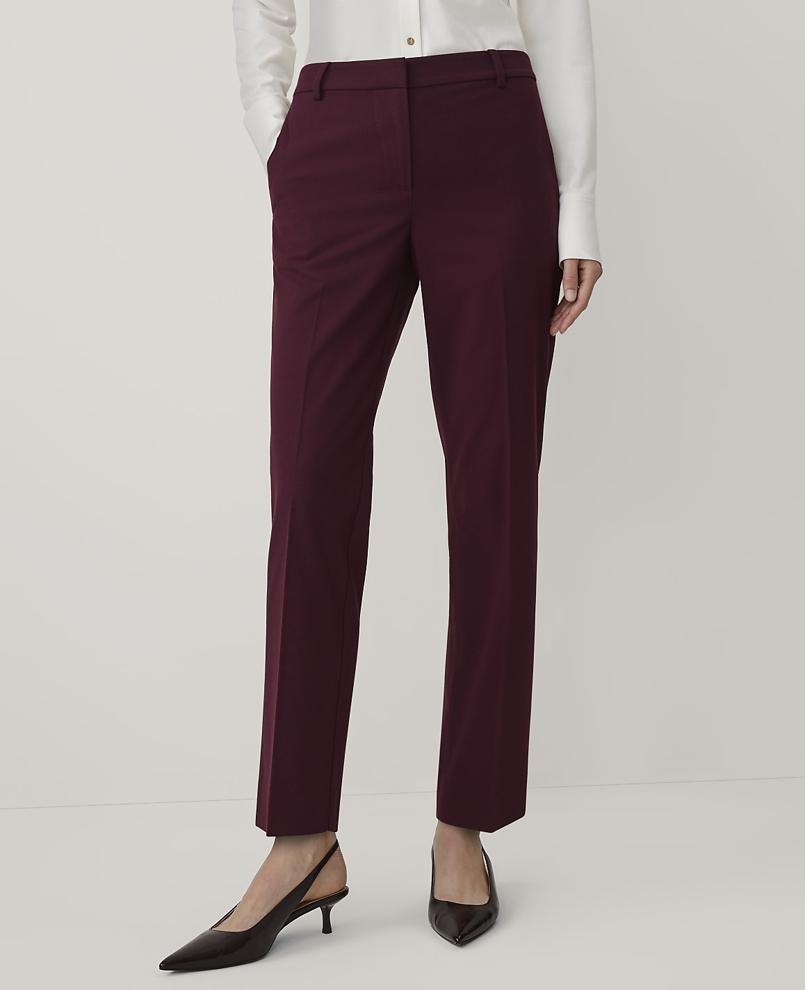 A person wearing a white shirt, burgundy trousers, and dark pointed-toe heels stands against a plain white background.