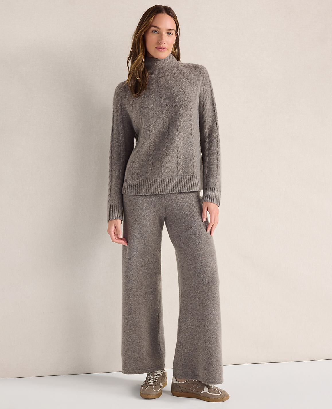 A woman wearing a grey cable-knit sweater and matching wide-leg trousers stands against a plain, neutral-colored background.