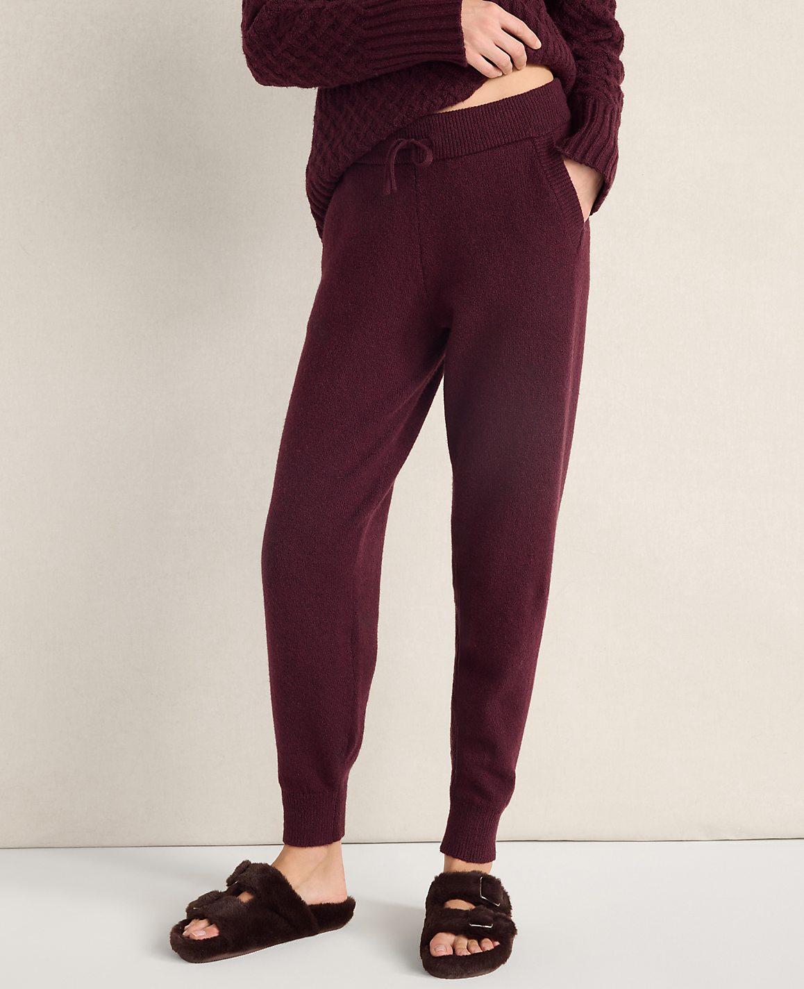 A person wears burgundy knit joggers and fuzzy brown slippers against a plain, light-colored wall.