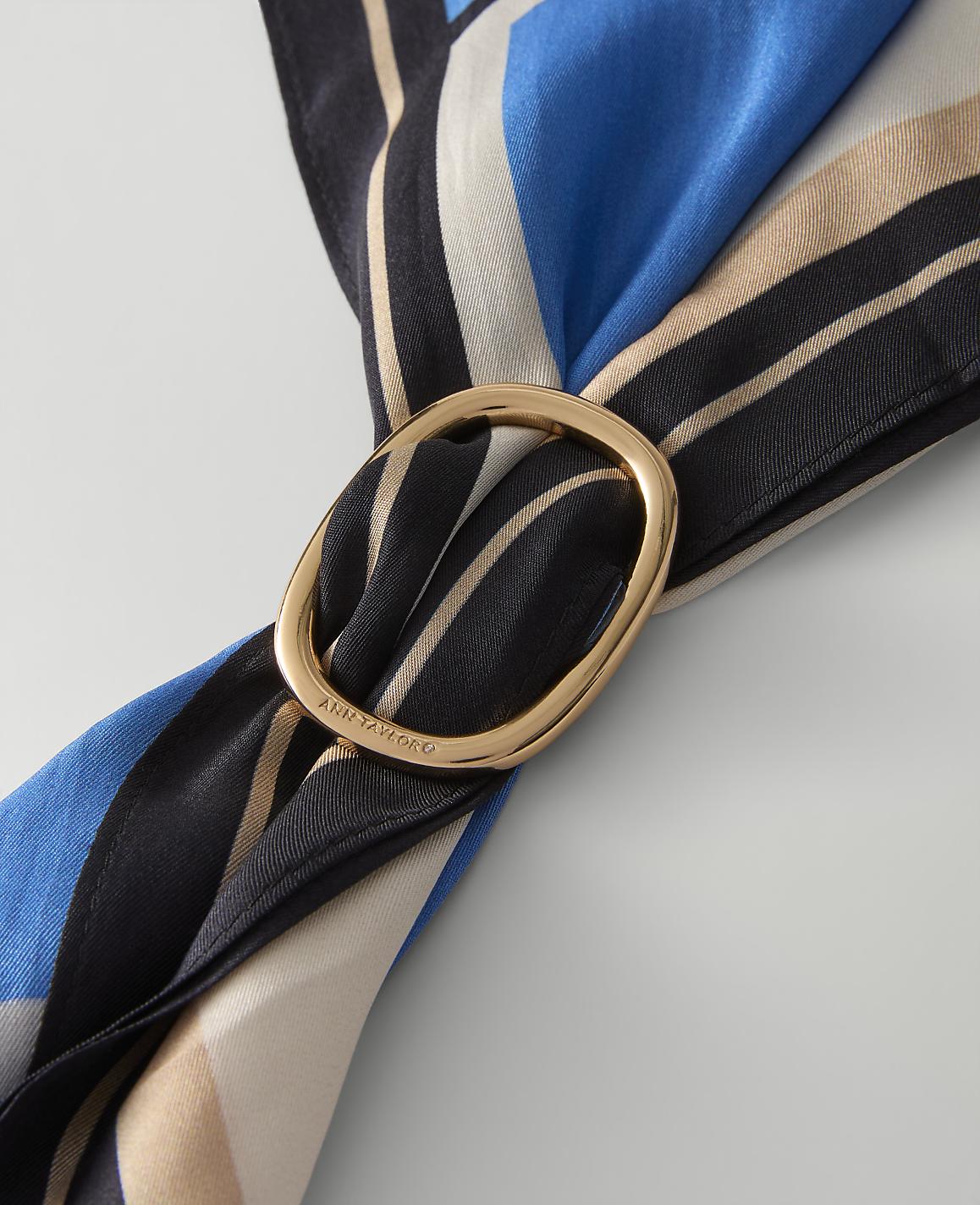 A blue, black, and beige patterned fabric is looped through a gold-toned oval buckle against a plain light gray background.
