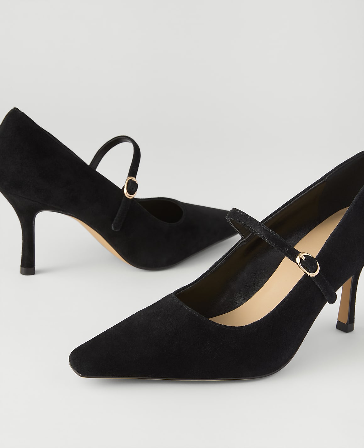 A pair of black suede pointed-toe pumps with ankle straps are positioned against a plain, light grey background.