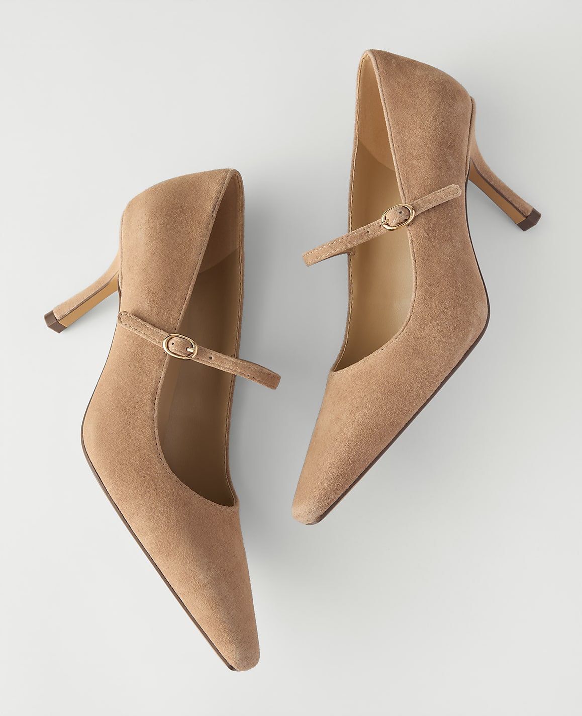 A pair of tan suede pointed-toe pumps with ankle straps rests on a plain white background.