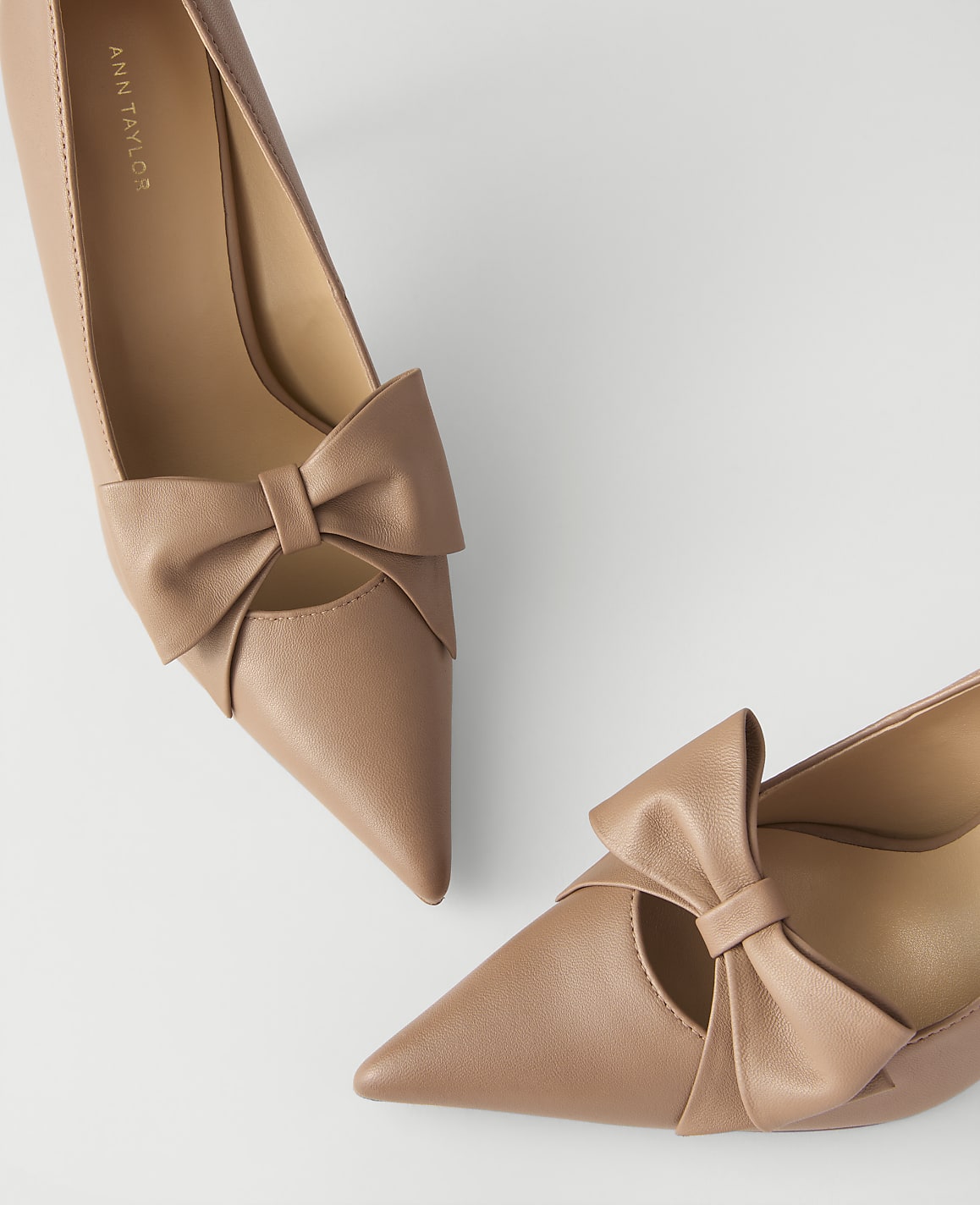 Two beige pointed-toe pumps with decorative bows are positioned against a plain white background.