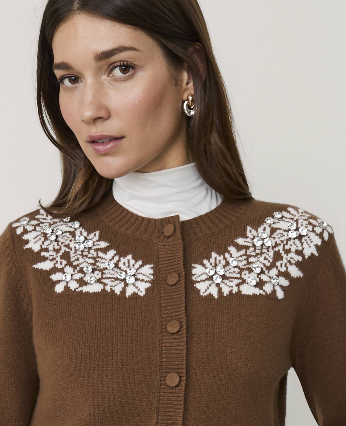A woman wearing a brown cardigan with white floral embellishments and a white turtleneck stands against a plain, light-colored background.