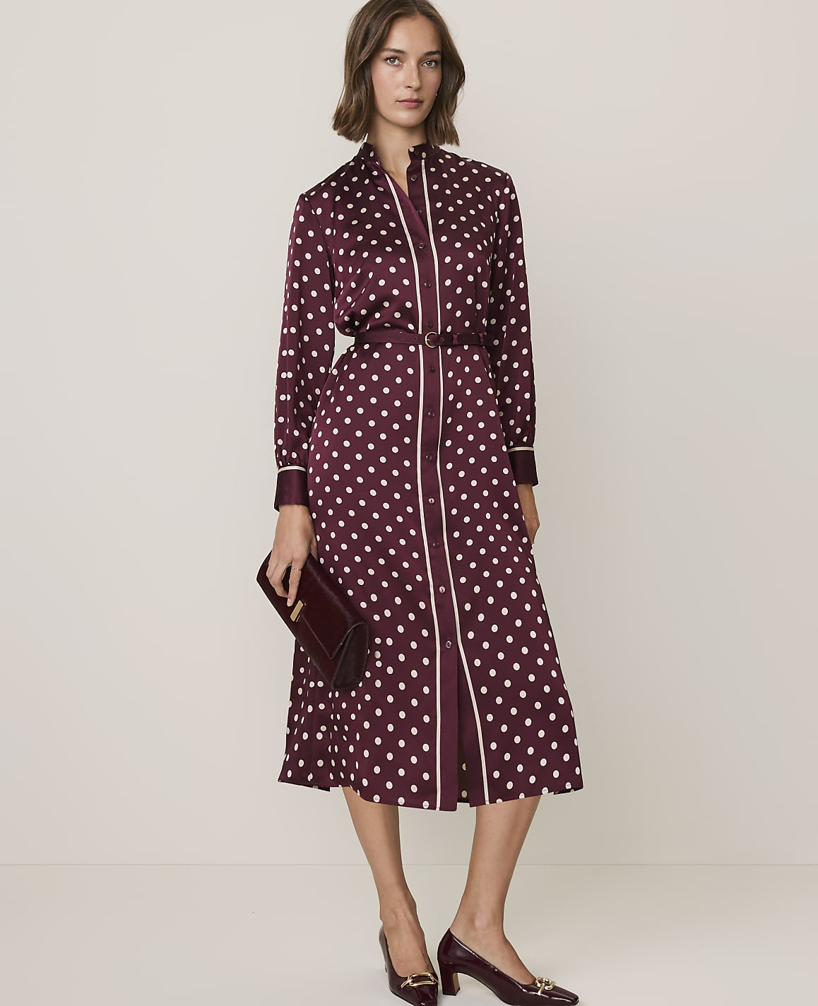 A woman in a burgundy polka dot dress with a pussy bow stands against a plain, light-colored background.