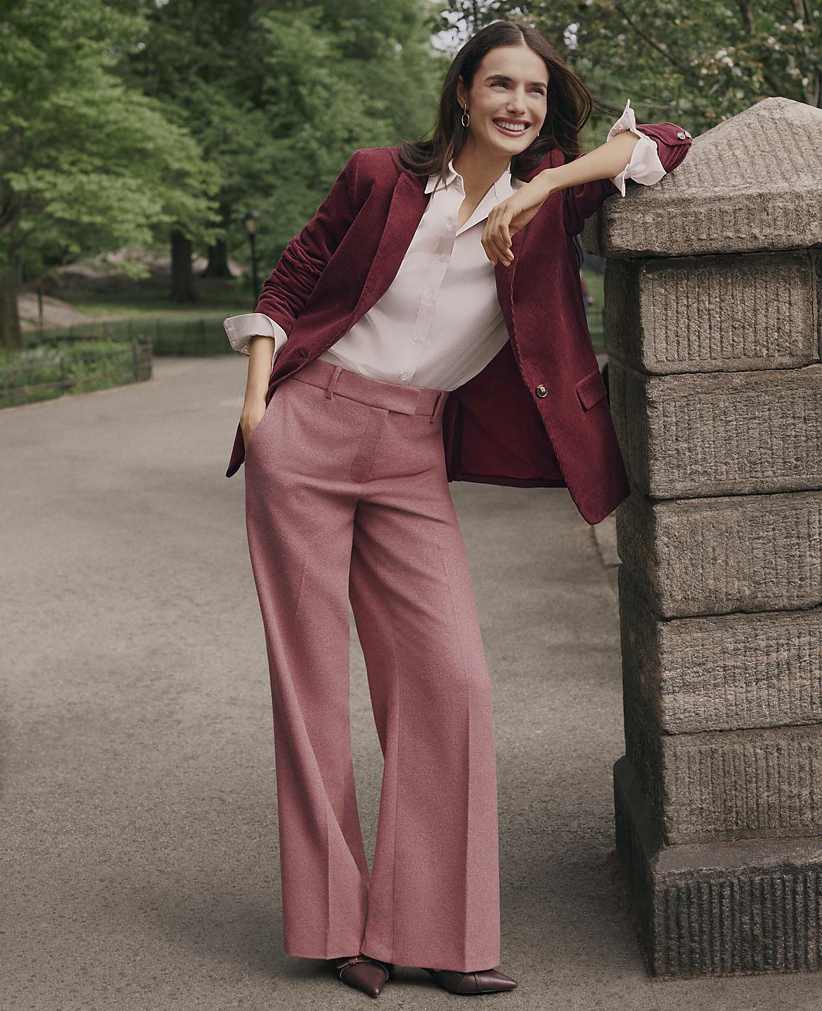 A smiling woman in a burgundy blazer and pink wide-leg trousers leans against a stone pillar on a paved path surrounded by lush green trees.