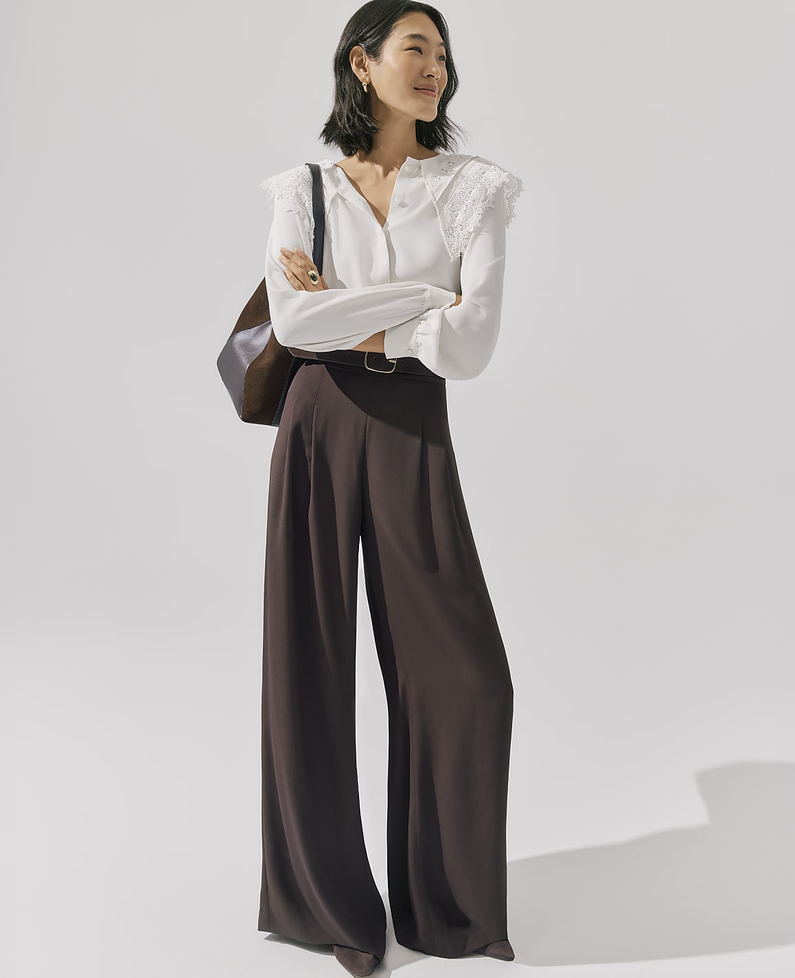 A woman wearing a white lace-detailed blouse and wide-leg brown trousers stands with her arms crossed against a plain, light-colored background.