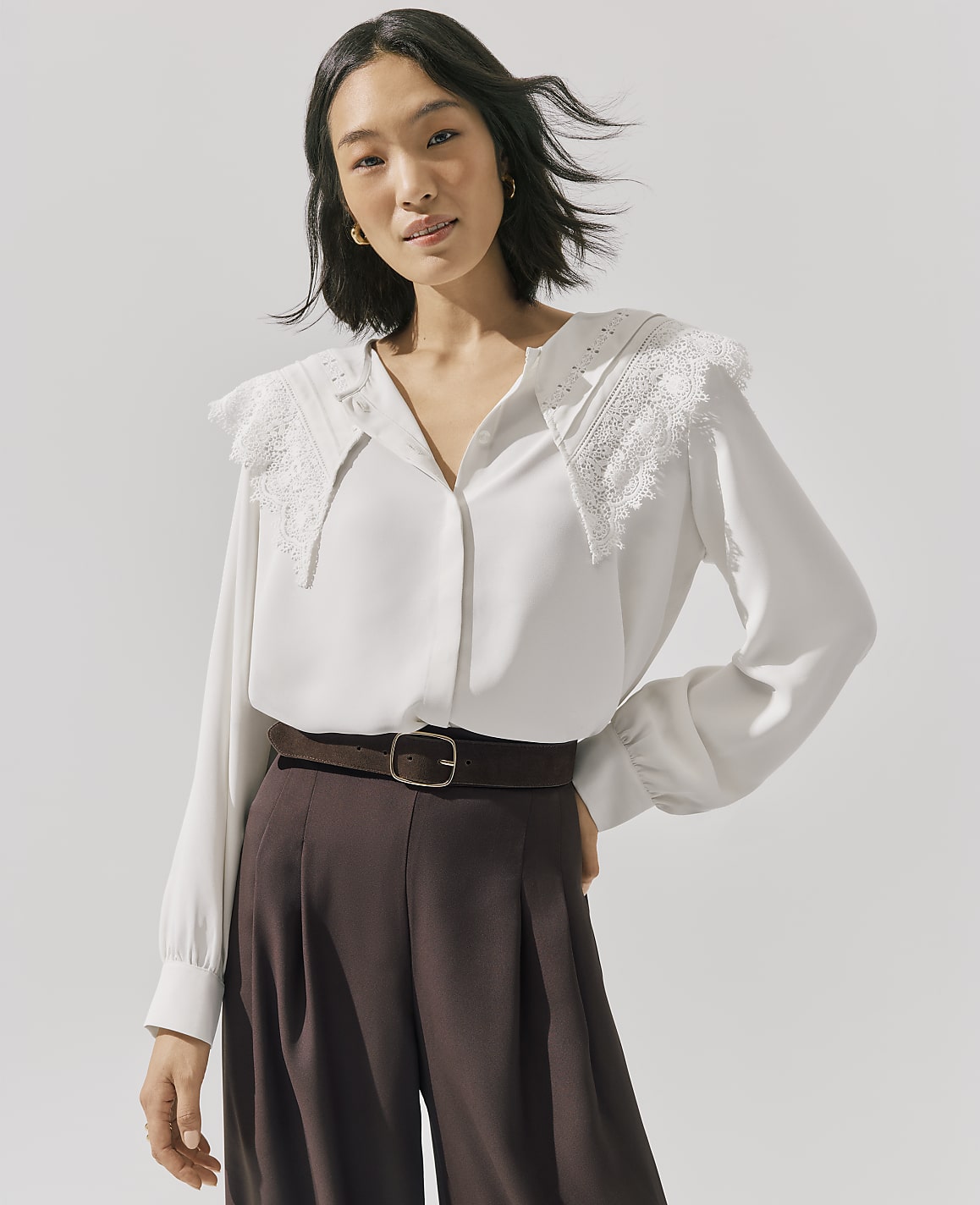 A woman wearing a white lace-collared blouse and brown trousers stands against a plain light gray background.