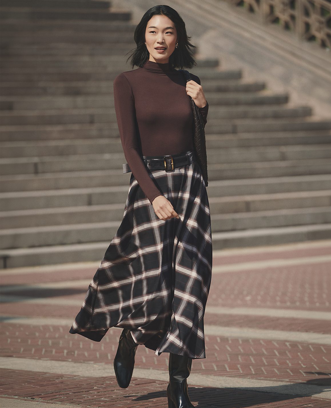 A woman wearing a brown turtleneck and a plaid skirt walks across a brick plaza in front of a large set of stone stairs.