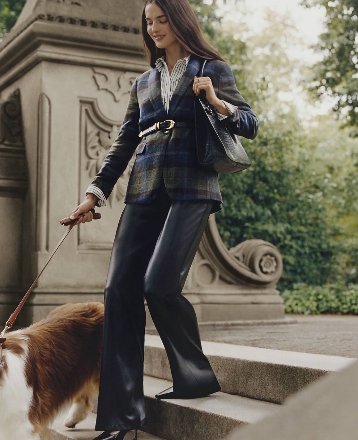 A woman in a plaid blazer and leather trousers walks a brown and white dog down stone steps against a backdrop of lush green foliage.