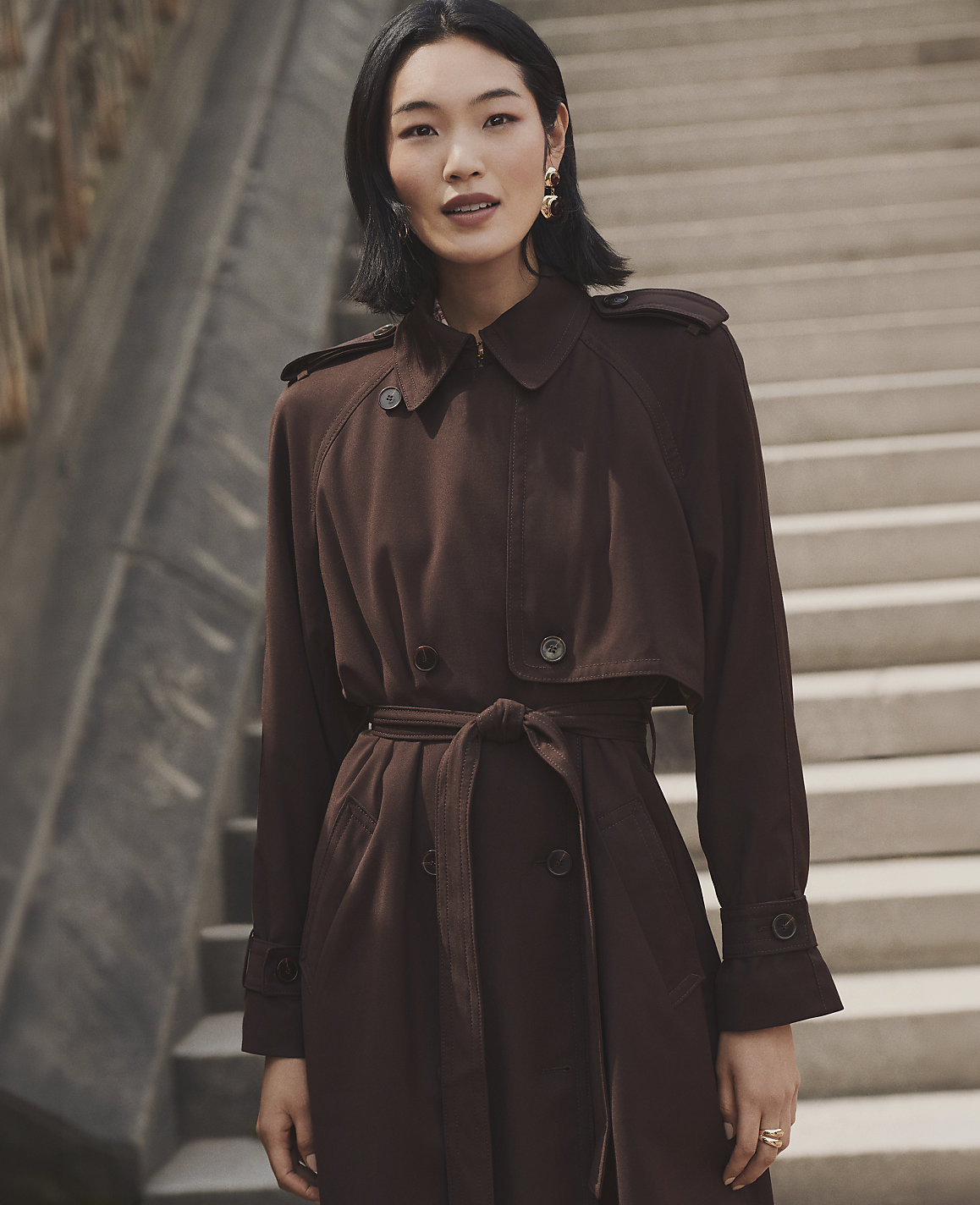 A woman wearing a brown belted trench coat stands in the foreground against a background of concrete stairs.