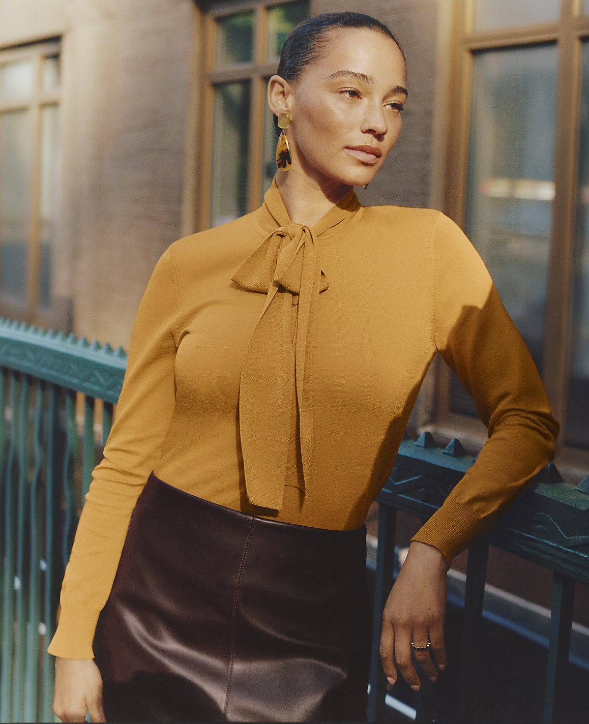 A woman wearing a mustard-yellow pussy-bow blouse and a dark leather skirt leans against a teal railing in front of a blurred city building.