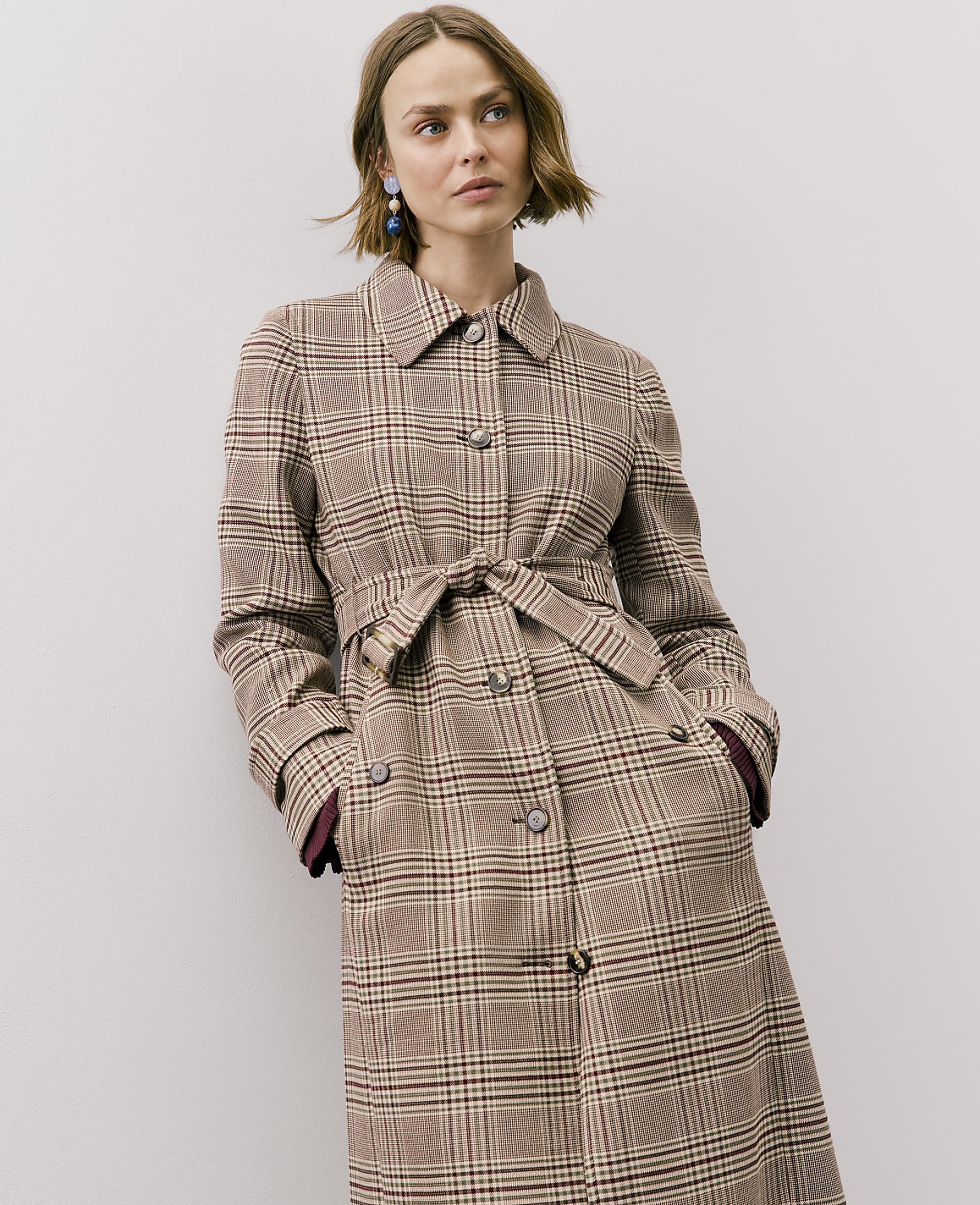 A woman wearing a brown plaid belted coat stands against a plain white background.