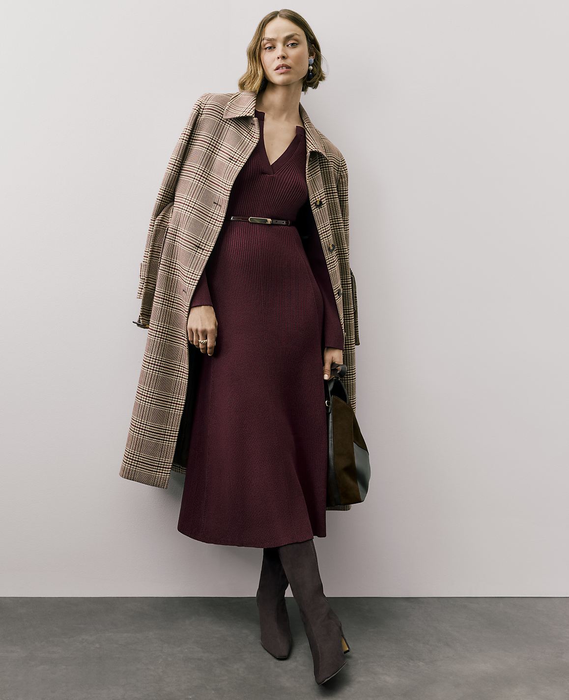 A woman wearing a burgundy ribbed dress, a plaid coat, and dark boots stands against a plain, light-colored wall and grey floor.