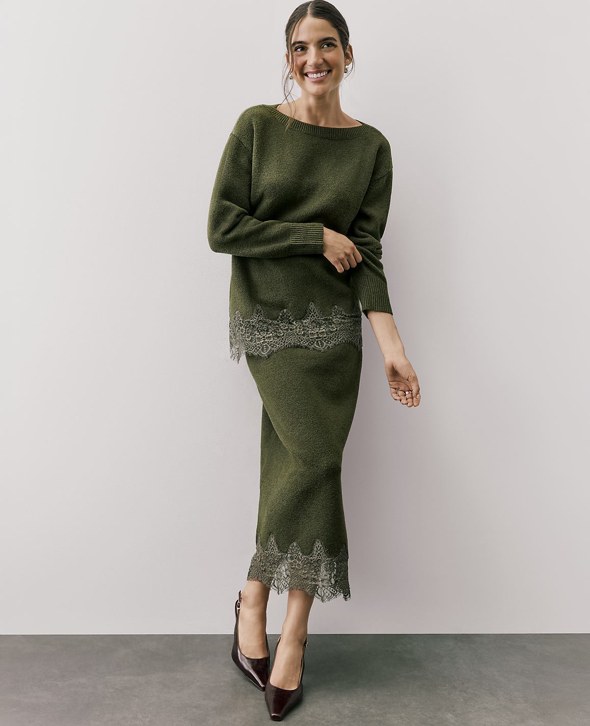 A smiling woman wears a green knit two-piece set with lace trim, standing against a plain white wall and grey floor.