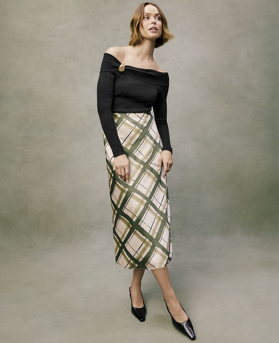 A woman wearing a black off-the-shoulder top and a plaid midi skirt stands against a muted green textured background.