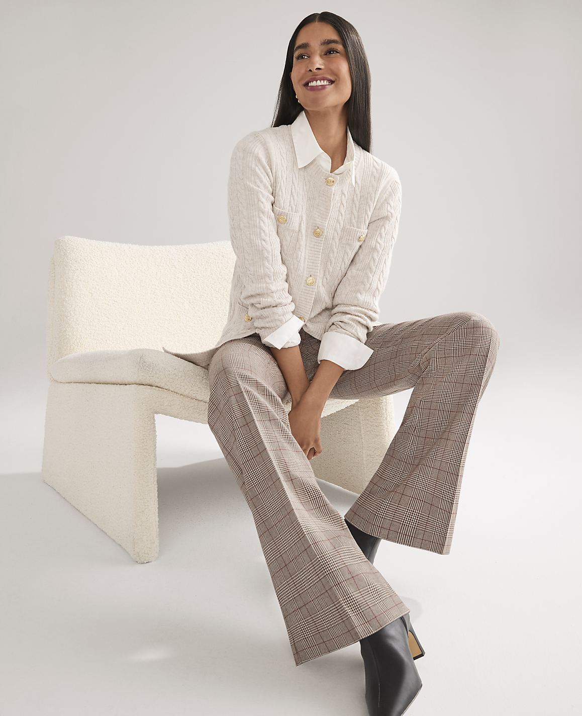 A smiling woman wearing a white cable-knit cardigan and plaid trousers sits on a white textured chair against a plain light gray background.
