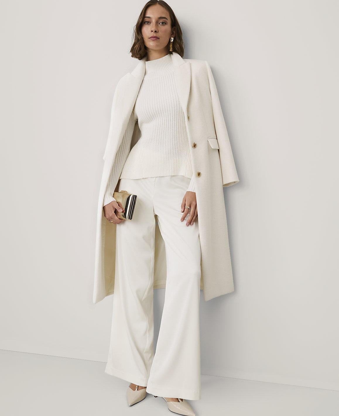 A woman wearing an all-white ensemble consisting of a ribbed sweater, wide-leg trousers, and a long coat stands against a plain white wall.
