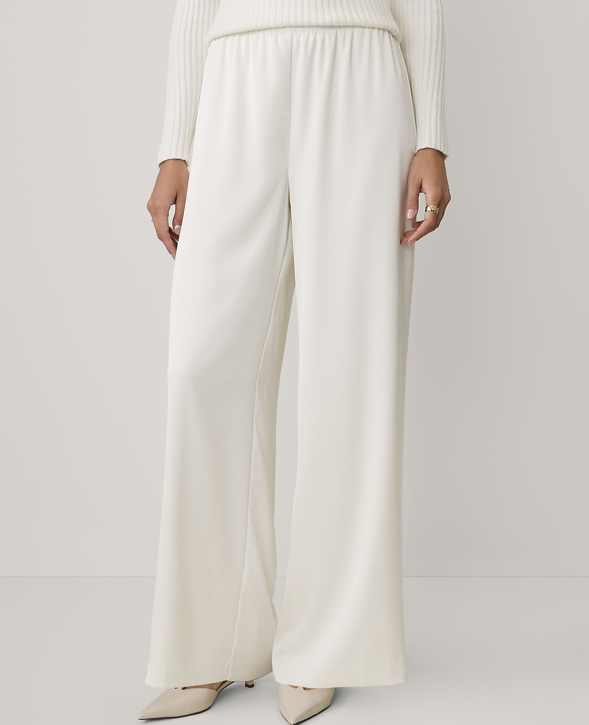A person wearing a white ribbed sweater and wide-leg white trousers stands against a plain, light-colored background.
