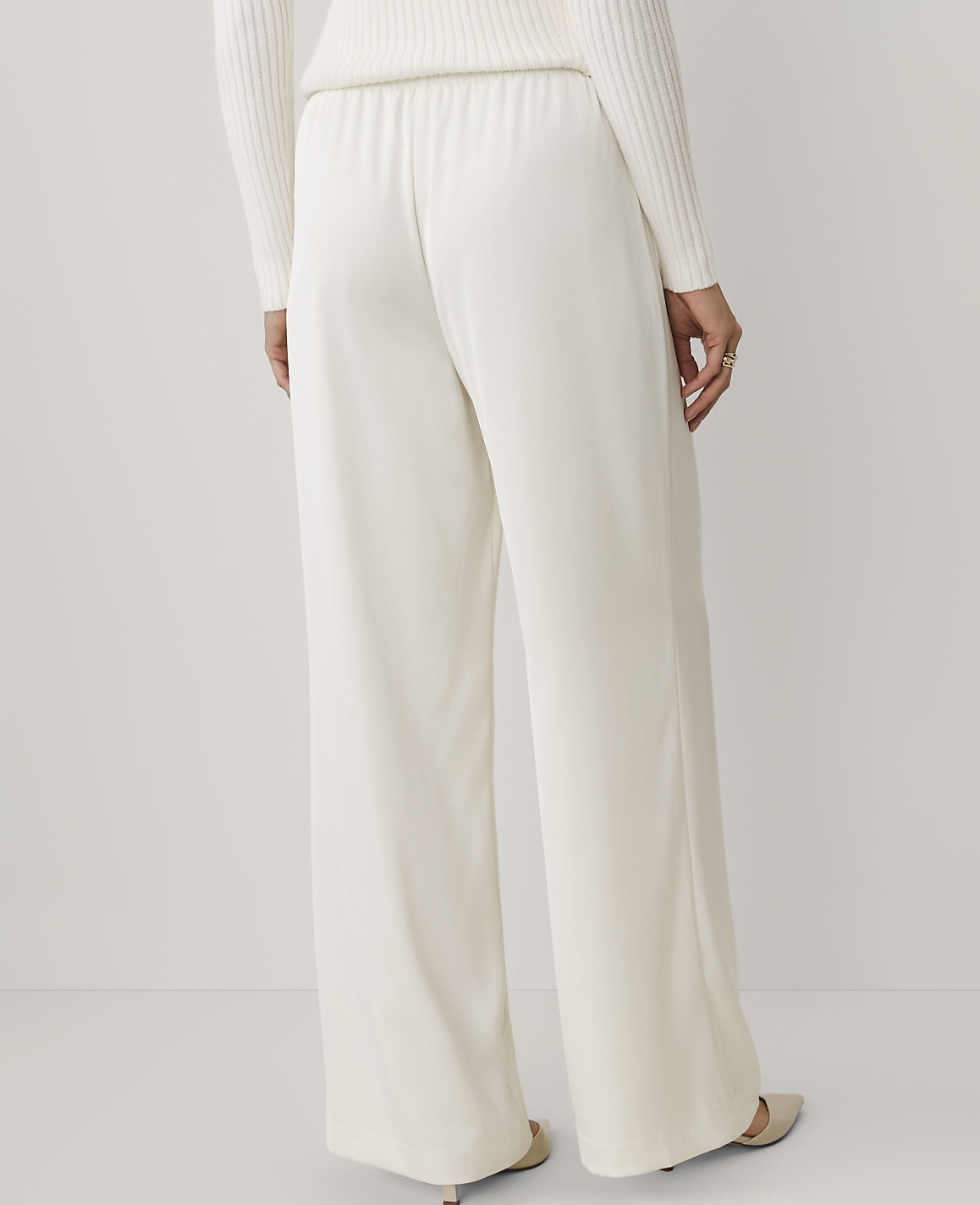 A person wears wide-leg white trousers and a white sweater against a plain, light-colored background.