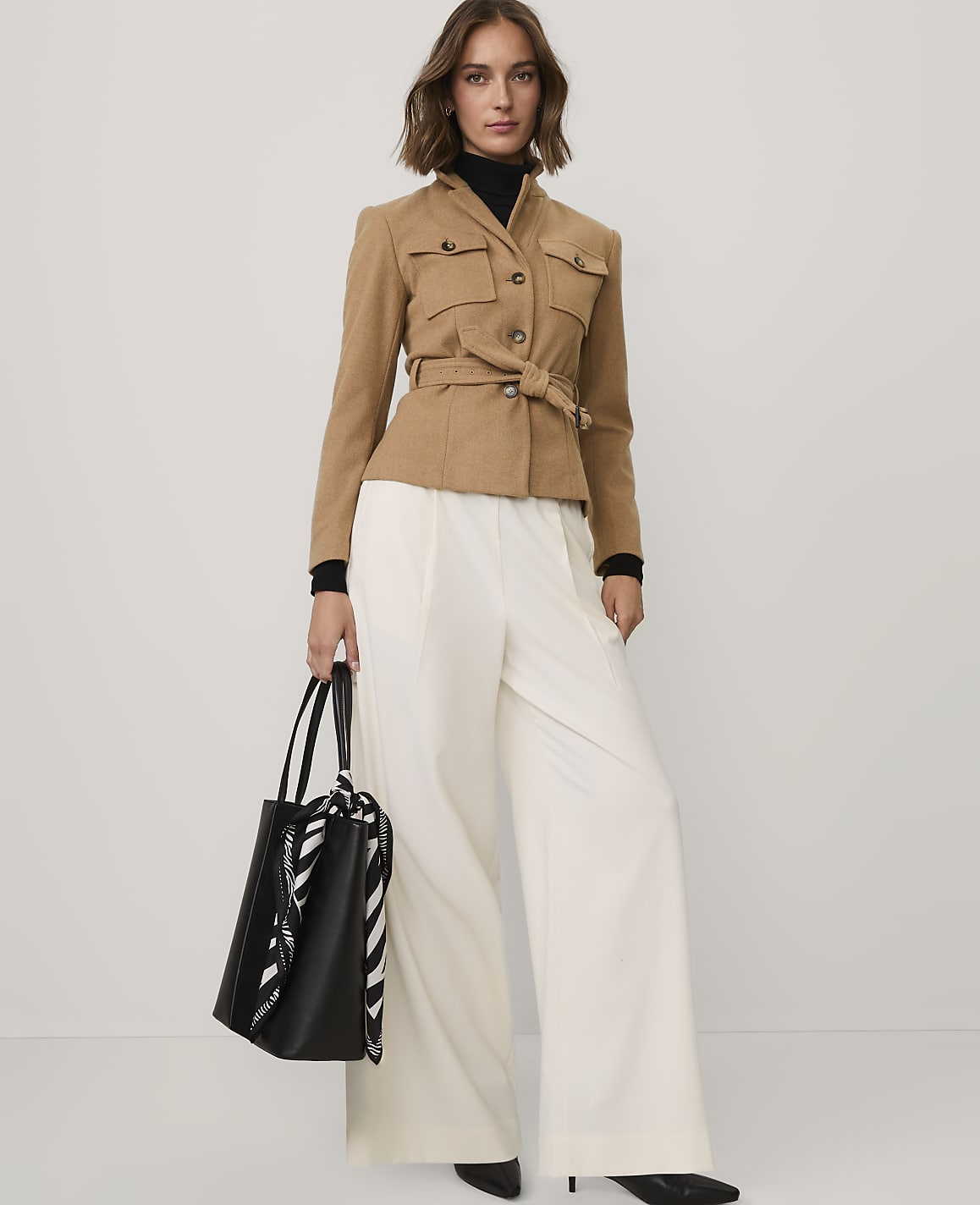 A woman wearing a tan belted jacket and wide-leg white trousers stands against a plain light gray background.
